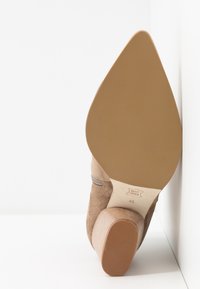 Ruskea mokkanahkainen korkokenkä, jossa on kartiomainen korko ja vaaleanbeige pohja. Sivussa on koristeellinen harmaa vetskari.