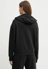 Donna con capelli castani che indossa un semplice cappuccio nero e pantaloni neri, mostrata di spalle su uno sfondo chiaro e neutro.