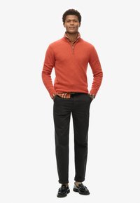 Pullover arancione con zip, indossato sopra una camicia a quadri, abbinato a pantaloni neri e mocassini neri, con le maniche arrotolate.