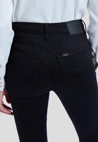 Isik kannab musta kitsaid teksapükse tagataskute, vööaugude ja väikese 'Lee' brändisildiga paremal taskul.