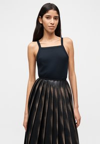 Γυναίκα με μακριά σκούρα μαλλιά που φοράει μαύρο τοπ με τετράγωνο ντεκολτέ και μαύρη ψηλόμεση φούστα με πιέτες και μπρονζέ ανταύγειες.