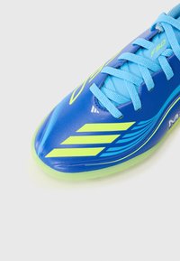Zapato de fútbol azul con un acabado brillante, que presenta franjas y acentos amarillos, cordones texturizados y una suela verde claro.