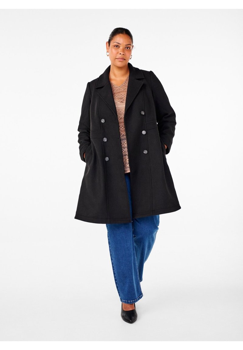 Vrouw staand met handen in de zakken, draagt een zwarte dubbelgeknoopte jas, een beige gebreide top, blauwe spijkerbroek en zwarte puntige schoenen op een witte achtergrond.