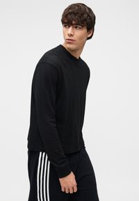 Μαύρο μπλουζάκι μακρυμάνικο από απαλό βαμβάκι με στρογγυλή λαιμόκοψη· το μοντέλο φοράει μαύρο παντελόνι με λευκές ρίγες στο πλάι.