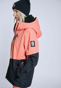 Vandafvisende jakke med en livlig koral top og sort bund, med hætte, lynlåslommer og et logo-patch på ærmet.