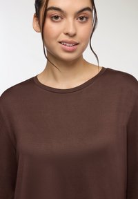 Brązowa bluzka z długim rękawem, z okrągłym dekoltem i gładką teksturą. Projekt jest prosty, bez wyraźnych wzorów czy akcentów.