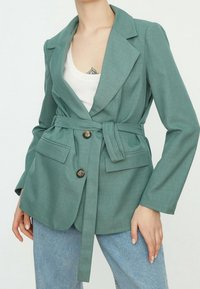 Teal blazer with a belted waist, dual button closure, two front pockets, and notched lapels, paired with a white tank top and blue jeans.