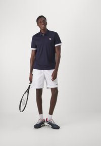 Marinblå polo med hvite detaljer, hvite shorts og gråblå tennissko. Holder en svart tennisracket og står på en lys bakgrunn.