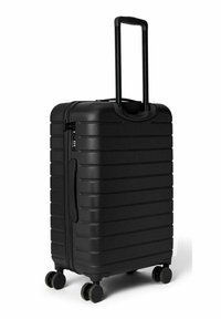 Valise rigide noire à surface texturée, avec des rainures verticales, une poignée extensible et quatre roues pour la mobilité. Dispose d'une poignée latérale.