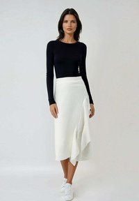 Un haut noir à manches longues associé à une jupe midi blanche côtelée présentant un détail drapé sur le côté. Le mannequin porte des baskets blanches.