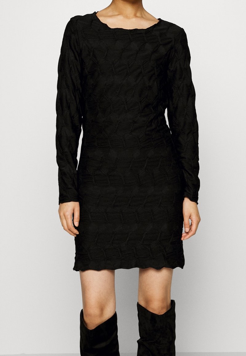 Femme portant une robe noire ajustée texturée à manches longues et des bottes noires montant jusqu'aux genoux, debout devant un fond blanc uni.
