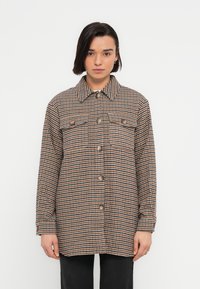 Καρό πουκάμισο-ζακέτα σε καφέ, κρεμ και μαύρο. Διαθέτει γιακά, κουμπιά μπροστά και δύο τσέπες στήθους. Κατασκευασμένο από υφασμάτινη υφή.