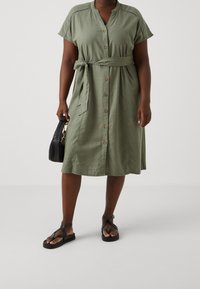 Oliwkowa sukienka z krótkim rękawem, zapinana na guziki, z detalem w postaci wiązania w talii, bocznymi kieszeniami i luźnym krojem. Połączona z czarnymi sandałami na krzyż i torebką.