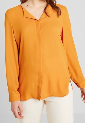 Žena oblečená v hořčicově žluté blůze s dlouhými rukávy a knoflíky, doplněná krémovými kalhotami, na jednobarevném pozadí.