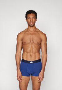 Boxers de algodón azules con una cinturilla negra que presenta un logo blanco, llevados por una persona de complexión muscular sobre un fondo sencillo.