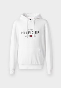 Hvit hettegenser med frontlomme, som har «Tommy HILFIGER»-logo i svart og et lite design med rød og blå.flagg. Myk stofftekstur.
