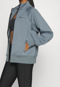 Ljusblå zip-jacka med hög krage, med sidfickor och en Champion-logga på bröstet, tillverkad av ett slätt, strukturerat tyg.