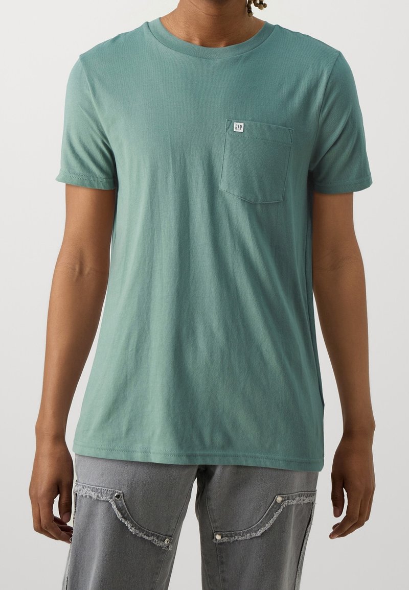 Personne portant un t-shirt vert uni à manches courtes avec une poche avant et une étiquette, associé à un pantalon cargo gris aux bords de poche effilochés.