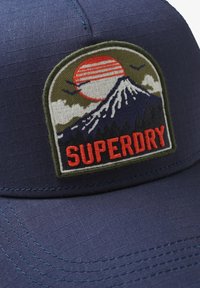 Marineblauwe pet met een geborduurde patch waarop een zon, berg en bomen zijn afgebeeld. De patch heeft groene, rode en witte details met de tekst "SUPERDRY".