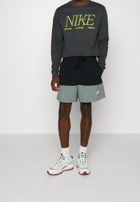 Dunkelgraues Nike-Shirt mit langen Ärmeln und gelbem Logo, kombiniert mit schwarz-grauen Shorts. Weiße Nike-Sneaker mit roten Akzenten und weißen Socken.