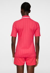 Różowa koszulka polo sportowa z krótkimi rękawami, pionową żebrowaną strukturą, kontrastowym białym kołnierzykiem i dopasowanymi szortami, noszona przez osobę widzianą z tyłu.