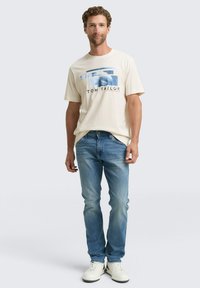 Hombre de pie con una camiseta gráfica color crema, jeans azules y zapatillas blancas sobre un fondo liso gris claro.