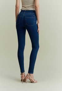 Jeans skinny en denim bleu foncé, dotés de cinq poches et d'une couture subtile, associés à des sandales à talons hauts nudes sur un fond clair.