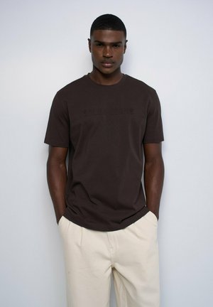 Joven con el pelo corto, vestido con una camiseta marrón oscuro y pantalones beige claros, de pie frente a un fondo claro y liso.