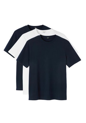 Jeune homme aux cheveux blonds courts portant un t-shirt bleu marine uni, un pantalon clair rayé et des tatouages visibles sur les deux avant-bras.