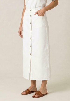 Femme portant une jupe midi blanche boutonnée sur le devant avec des poches et des sandales plates ouvertes marron, debout sur un fond neutre.