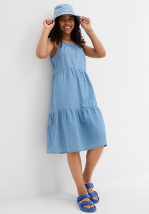 Abito in denim azzurro chiaro con un design a strati, spalline sottili e vita arricciata. Abbinato a un cappello a secchiello coordinato e sandali blu.