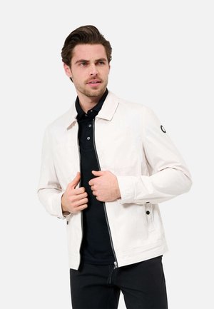 Hellbeige Jacke mit einem kragenbetonten Design, Reißverschluss, zwei Taschen und einem schwarzen Logo-Akzent am linken Ärmel. Getragen über einem schwarzen Shirt.