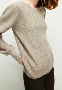 Beige Strickpullover mit rundem Halsausschnitt, strukturiert mit goldenen Fäden, langen Ärmeln und gerippten Bündchen und Saum. Getragen mit dunklen Hosen.