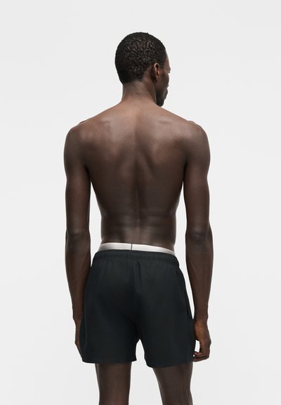 Άνδρας με σκουρόχρωμο δέρμα και κοντά μαλλιά, όρθιος με την πλάτη στραμμένη προς την κάμερα, φορώντας μαύρο σορτς με λευκή μέση, μπροστά σε μονόχρωμο γκρι φόντο.