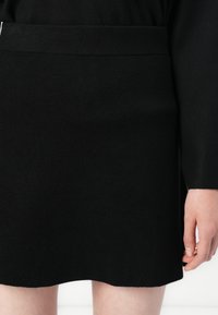 Mujer con falda negra lisa hasta la rodilla y top negro de manga larga, con la mano apoyada a un lado.