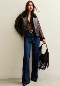 Bruine leren cropped jas met een bontkraag, gecombineerd met donkerblauwe flared jeans. Houdt een zwarte franje handtas vast, met zwarte haklaarzen.