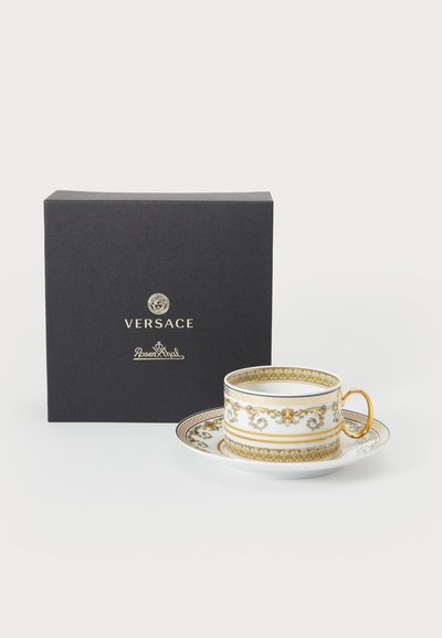 Πορσελάνινη κούπα τσαγιού με πιατάκι και χρυσά σχέδια, με διακοσμητικό περίγραμμα και χρυσή λαβή, δίπλα σε μαύρο κουτί δώρου Versace.