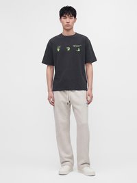 Young man wearing black Off-White t-shirt with green hand graphics and white drawstring sweatpants and white sneakers standing against a plain background.
