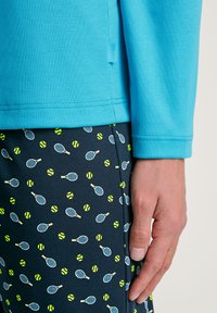 Blaues Langarmhemd mit einem Etikett, kombiniert mit dunklen Shorts, die ein Muster aus Tennisschlägern und gelben Tennisbällen aufweisen.