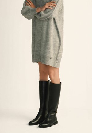 Osoba w szarej dzianinowej sukience swetrowej i czarnych skórzanych botkach za kolano, stojąca z założonymi rękami.