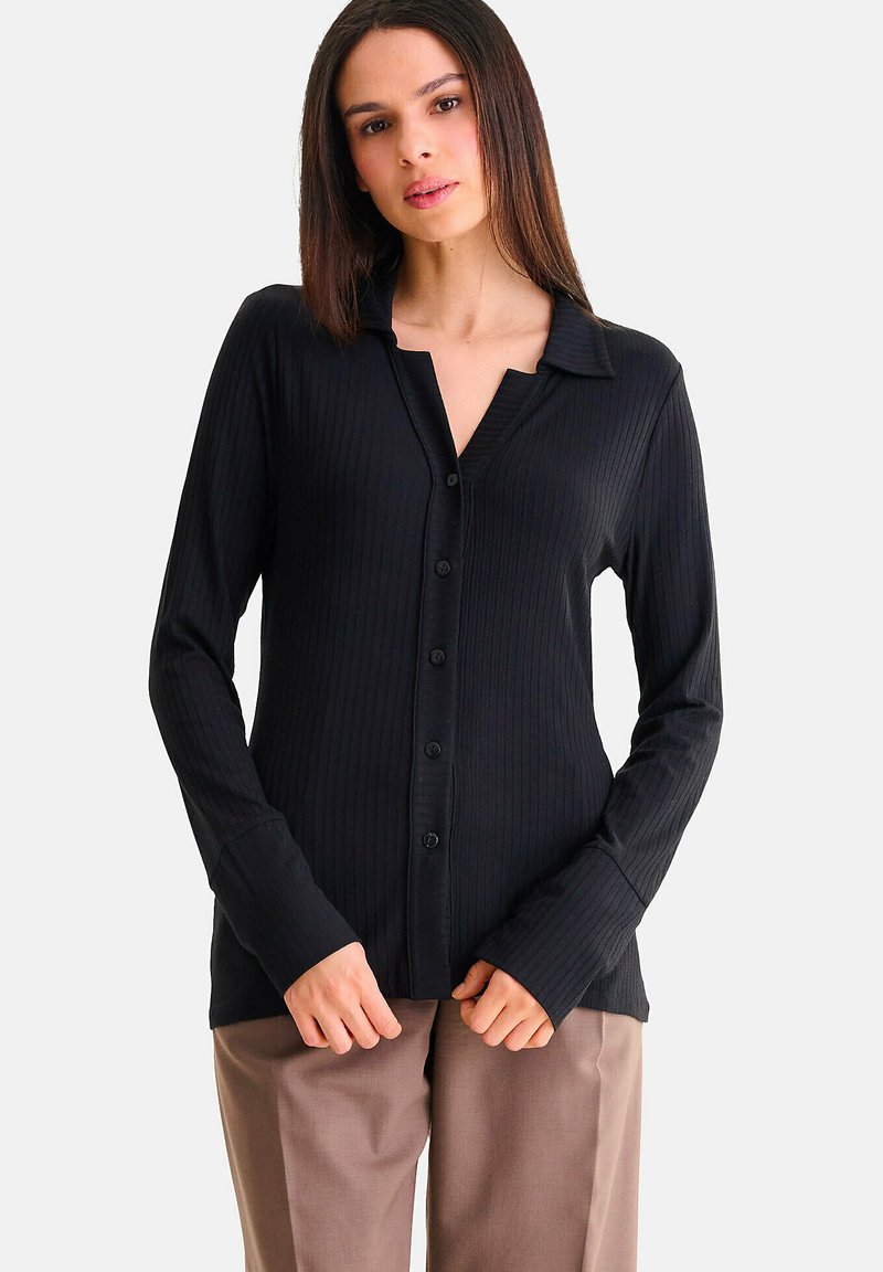 Zwarte geribbelde langemouwen shirt met een knoopsluiting aan de voorkant en een kraag, met een V-halslijn en aansluitende manchetten, gecombineerd met een bruine broek.
