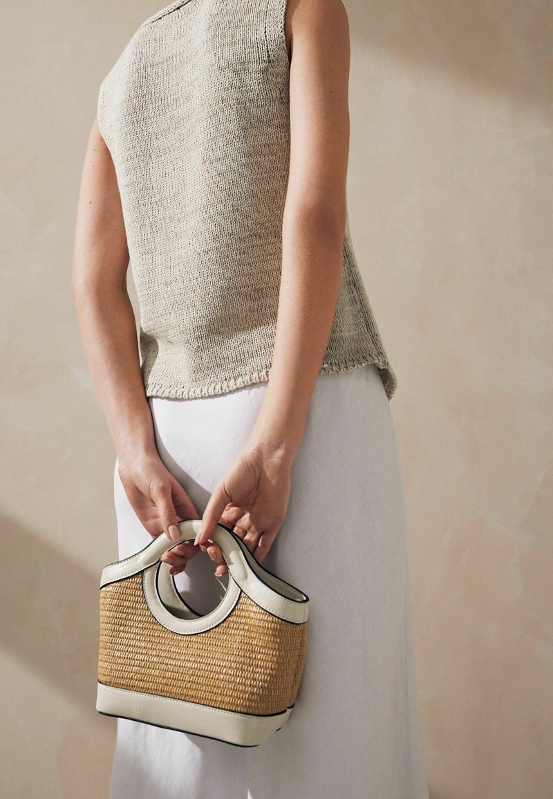 Femme en haut sans manches en maille beige et jupe blanche tenant un petit sac à main tressé beige et blanc derrière son dos.