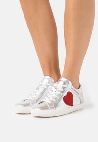 Zilverkleurige leren sneakers met een rood hartpatch en witte veters. Heeft een gladde textuur en een rubberen zool. Geaccentueerd met merknaamstiksels.