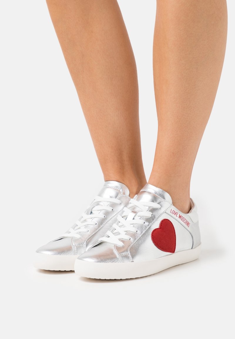 Zilverkleurige leren sneakers met een rood hartpatch en witte veters. Heeft een gladde textuur en een rubberen zool. Geaccentueerd met merknaamstiksels.