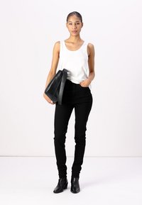 Woman with braided hair wearing white sleeveless top, black pants, black ankle boots, holding large black handbag, standing against plain background.