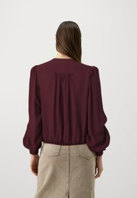 Blusa bordeaux con maniche a sbuffo, dettagli arricciati sul retro e una texture morbida. Abbinata a una gonna beige con tasche posteriori.