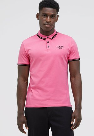 Mann trägt ein rosa Poloshirt mit schwarzem Besatz an Kragen und Ärmeln, schwarze Hose, steht vor einem schlichten hellgrauen Hintergrund.