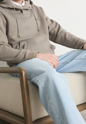 Homme assis en tailleur sur une chaise en bois beige avec coussin, portant un sweat à capuche taupe et un jean bleu clair sur fond blanc.