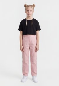 Fiatal lány szőke hajjal, két kontyban  és fonatokkal, fekete, rövid felsőt, rózsaszín nadrágot és fehér tornacipőt visel, előre nézve áll.