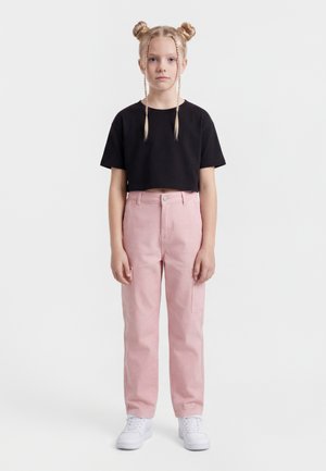 Mladé dievča s blond vlasmi v dvoch buchtičkách a vrkôčikoch, oblečená v čiernom krátkom tričku, ružových nohaviciach a bielych teniskách, stojí čelom dopredu.